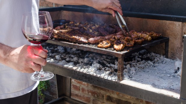 Man-cooking-a-traditional-Argentinian-barbecue-holding-a-glass-of-red-wine-on-his-hand.-Outdoor-coal-grill-with-cow-meat-ribs-and-sausages..jpeg